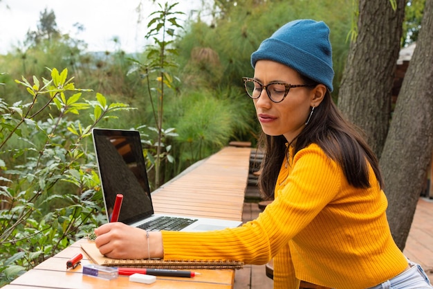 Jeune étudiante avec chapeau bleu et pull jaune écrivant dans un cahier à côté de son ordinateur portable assis dans un parc à l'extérieur