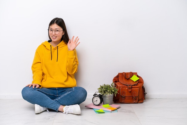 Jeune étudiante chinoise femme assise sur le sol isolé sur un mur blanc saluant avec la main avec une expression heureuse