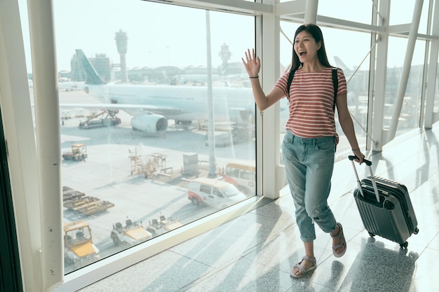 jeune étudiante chinoise de retour dans le pays de sa ville natale à l'aéroport de hong kong en agitant la main à la famille pour la chercher. collégienne joyeusement en vacances. belle femme marche dans le hall avec des bagages