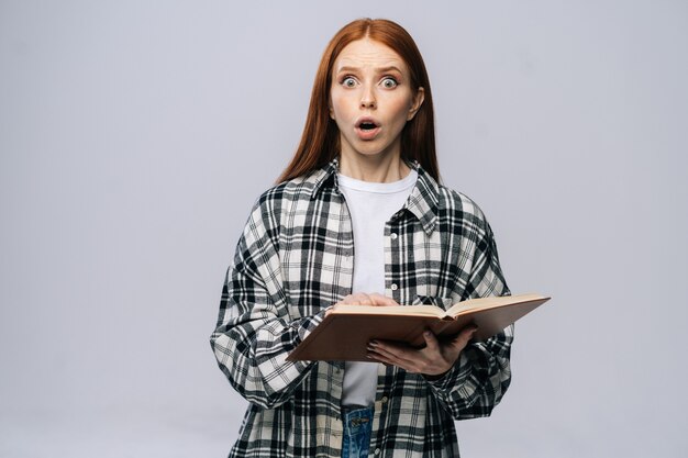 Jeune étudiante choquée avec la bouche ouverte tournant les pages du livre en lisant