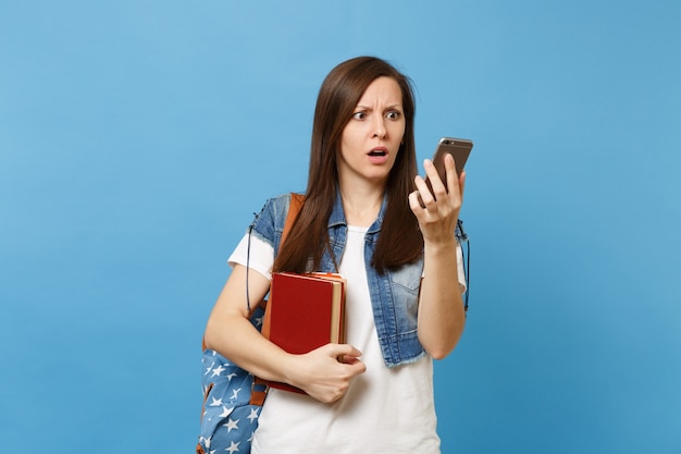 Jeune étudiante choquée et perplexe avec un sac à dos tenant des livres scolaires, regardant sur un téléphone portable après une conversation isolée sur fond bleu. Éducation dans le concept de collège universitaire secondaire.