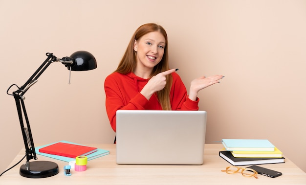 Jeune étudiante dans un lieu de travail avec un ordinateur portable tenant copyspace imaginaire sur la paume pour insérer une annonce