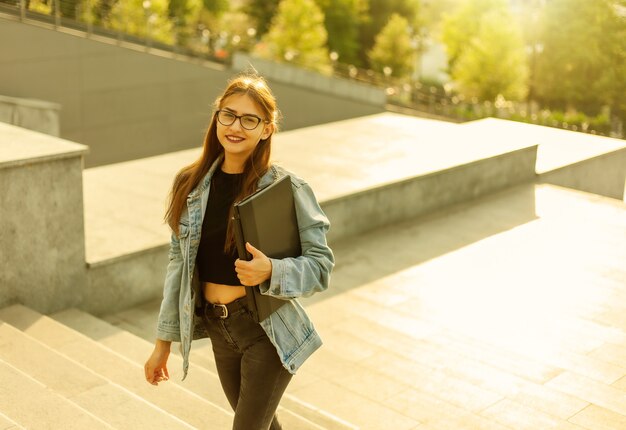 Une jeune étudiante dans une veste en jean et des lunettes monte les escaliers avec un ordinateur portable dans les mains dans la ville