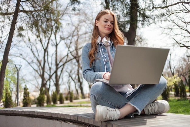 Une jeune étudiante est assise dans le parc sur un banc avec un ordinateur portable