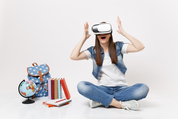 Jeune étudiante étonnée portant des lunettes de réalité virtuelle écartant les mains en s'asseyant près du globe, sac à dos, livres scolaires isolés sur mur blanc