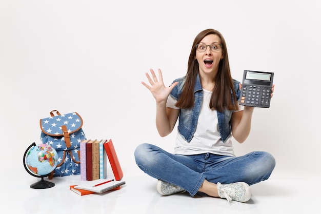 Jeune étudiante excitée choquée tenant une calculatrice écartant les mains apprenant les mathématiques assis près du globe, sac à dos, livres scolaires isolés