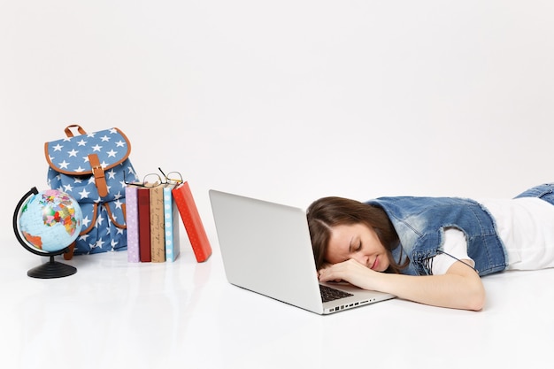 Jeune étudiante fatiguée en vêtements en jean dormant sur un ordinateur portable allongé près du sac à dos globe et des livres scolaires