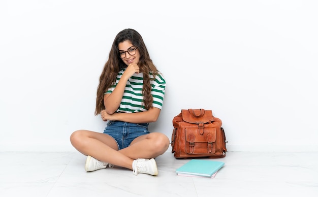 Jeune étudiante femme assise sur le sol isolé sur fond blanc avec des lunettes et souriant