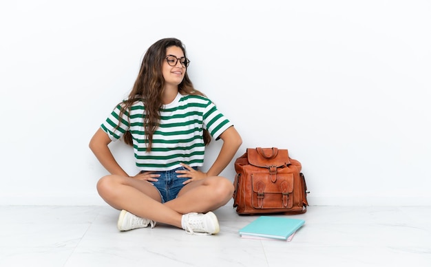 Jeune étudiante femme assise sur le sol isolé sur fond blanc posant avec les bras à la hanche et souriant