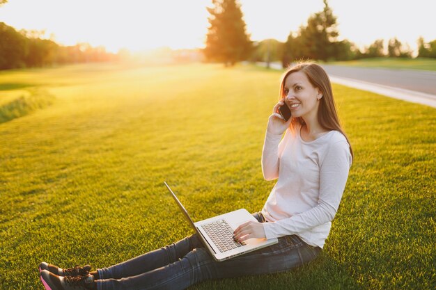 Jeune étudiante intelligente en vêtements décontractés. Femme parlant au téléphone portable, travaillant sur un ordinateur portable dans un parc de la ville sur une pelouse d'herbe verte ensoleillée à l'extérieur. Bureau mobile. Concept d'entreprise indépendant