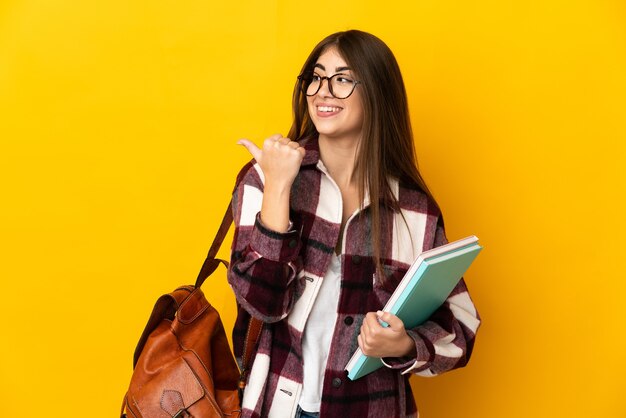 Jeune étudiante isolée sur un mur jaune pointant vers le côté pour présenter un produit