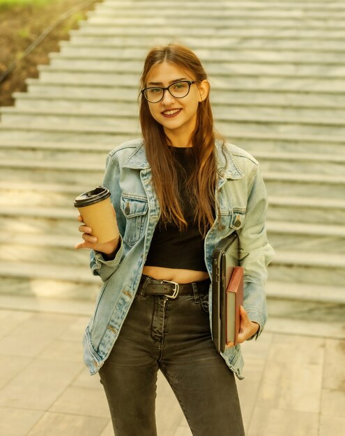 Jeune étudiante joyeuse dans une veste en jean et des lunettes descend les escaliers avec un ordinateur portable et une tasse de café dans les mains dans la ville
