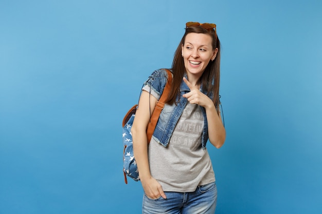 Jeune étudiante joyeuse et jolie en vêtements en jean avec sac à dos, lunettes coeur orange pointant l'index loin isolé sur fond bleu. L'éducation à l'université. Copiez l'espace pour la publicité.