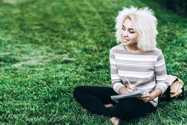 Une jeune étudiante joyeuse s&#39;assoit sur une herbe au campus