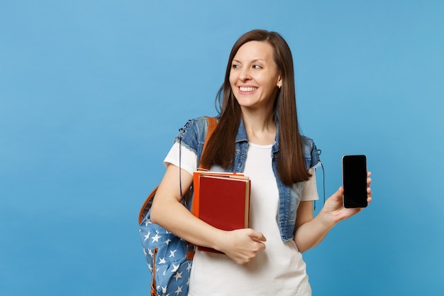 Jeune étudiante joyeuse en vêtements en denim avec sac à dos à côté de tenir des livres, téléphone portable avec écran vide noir blanc isolé sur fond bleu. Éducation au collège universitaire secondaire.