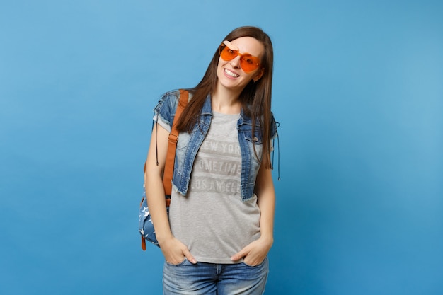 Jeune étudiante joyeuse en vêtements en denim avec sac à dos portant des lunettes coeur orange gardant les mains dans les poches isolées sur fond bleu. L'éducation à l'université. Copiez l'espace pour la publicité.