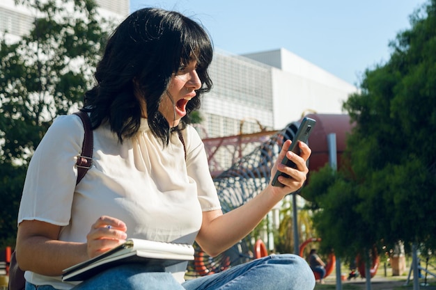 Jeune étudiante latine très surprise avec la bouche ouverte regardant le téléphone à l'extérieur