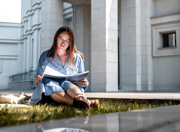 Jeune étudiante en lunettes assise sur l'herbe près du bâtiment de l'université et apprenant le livre de lecture f...