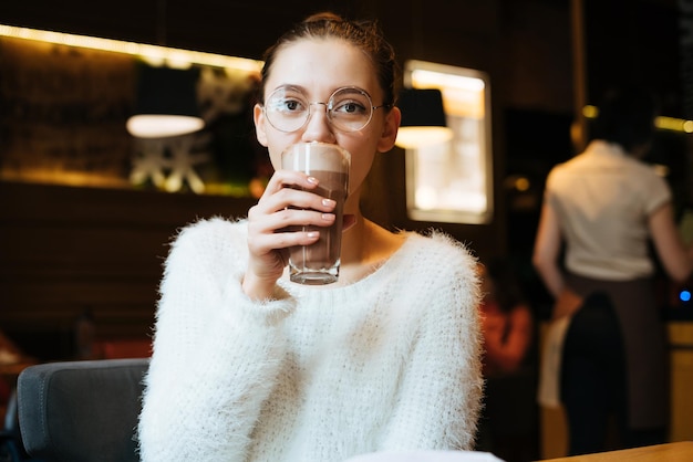Une jeune étudiante à lunettes et une veste blanche boit un délicieux café sucré dans un café