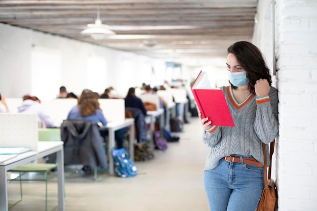 Jeune étudiante avec masque facial lisant un livre à l'intérieur d'une salle de classe Cours présentiels à l'université Espace pour le texte