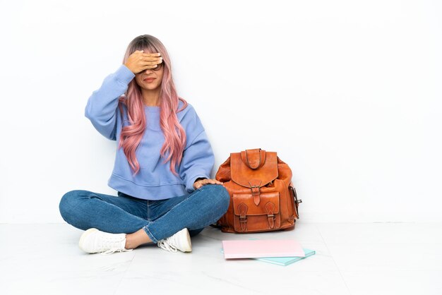 Jeune étudiante métisse femme aux cheveux roses assis sur le sol isolé sur fond blanc couvrant les yeux à la main. Je ne veux pas voir quelque chose