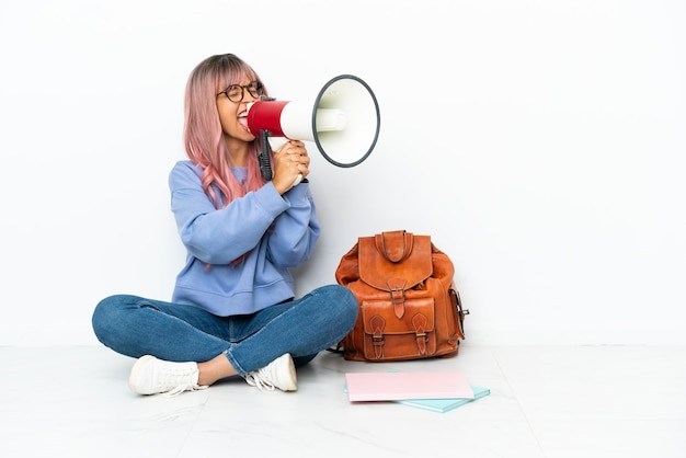 Jeune étudiante métisse femme aux cheveux roses assis sur le sol isolé sur fond blanc criant à travers un mégaphone