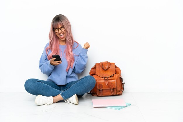 Jeune étudiante métisse femme aux cheveux roses assis sur le sol isolé sur fond blanc avec téléphone en position de victoire