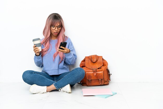 Jeune étudiante métisse femme aux cheveux roses assis sur le sol isolé sur fond blanc tenant du café à emporter et un mobile