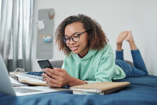Jeune étudiante métisse souriante en jeans et sweat à capuche défilant dans un smartphone en position allongée sur le lit devant un ordinateur portable dans sa chambre