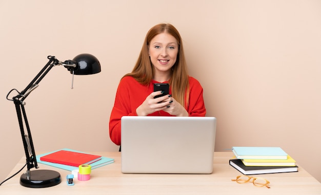 Jeune étudiante en milieu de travail avec un ordinateur portable en envoyant un message avec le mobile