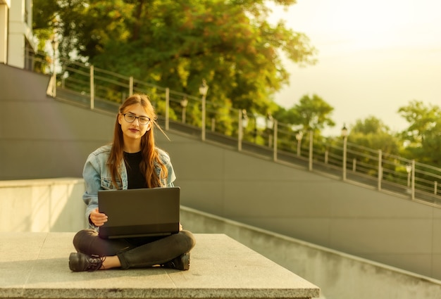 Jeune étudiante moderne dans une veste en jean assis sur les escaliers avec ordinateur portable. Apprentissage à distance. Concept de jeunesse moderne.