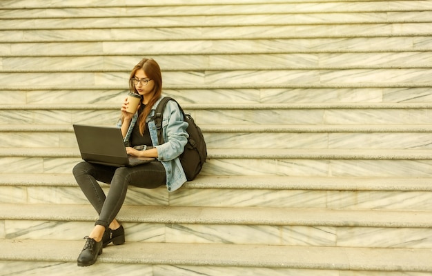 Jeune étudiante moderne dans une veste en jean assis sur les escaliers avec ordinateur portable. Apprentissage à distance. Concept de jeunesse moderne.