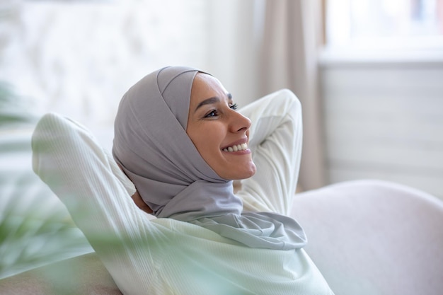 Jeune étudiante musulmane en hijab assise sur un canapé à la maison les mains derrière la tête et se reposant en souriant