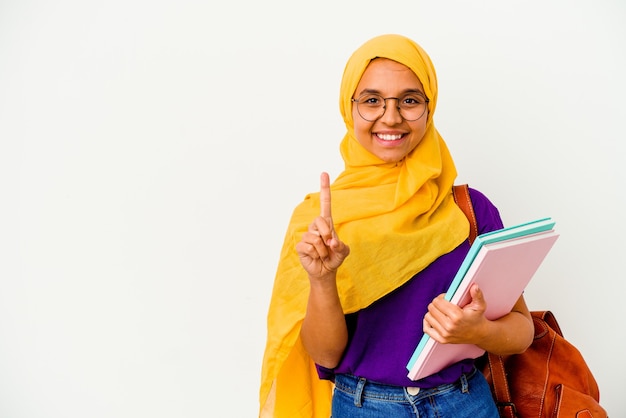 Jeune étudiante musulmane portant un hijab isolé sur un mur blanc montrant le numéro un avec le doigt.
