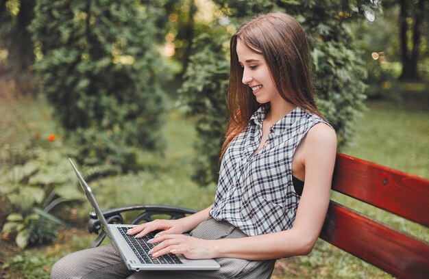 Jeune étudiante avec un ordinateur portable assis sur un banc dans le parc et en tapant sur le clavier