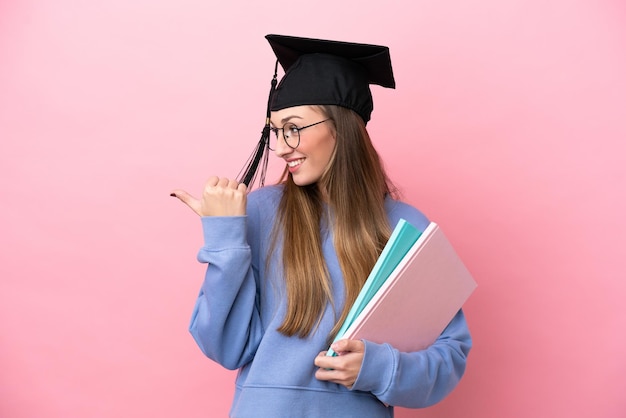 Jeune étudiante portant un chapeau de diplômé isolé sur fond rose pointant vers le côté pour présenter un produit