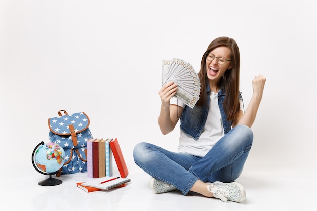 Jeune étudiante ravie tenant un paquet de dollars, de l'argent en espèces fait le geste du gagnant, dites oui près des livres de sac à dos globe isolés