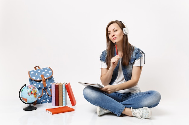 Jeune étudiante réfléchie dans les écouteurs écouter de la musique en pensant tenant un cahier, un crayon près du globe, des livres d'école de sac à dos isolés sur un mur blanc