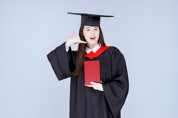 Jeune étudiante en robe académique montrant un livre rouge à la caméra. photo de haute qualité