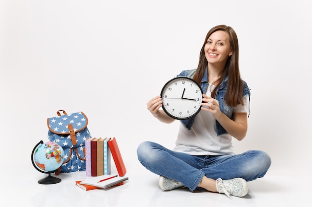Jeune étudiante souriante et agréable dans des vêtements en denim tenant un réveil assis près du globe, sac à dos, livres scolaires isolés