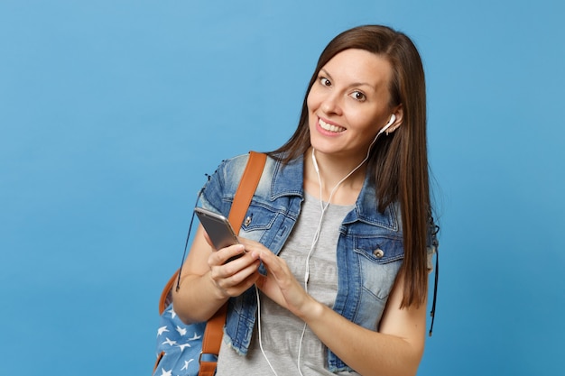 Jeune étudiante souriante en vêtements en denim avec sac à dos et écouteurs écoutant de la musique tenant à l'aide d'un téléphone portable isolé sur fond bleu. L'éducation au collège. Copiez l'espace pour la publicité.