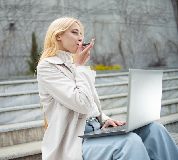 Une jeune étudiante utilise un ordinateur portable assis sur un banc et fume une cigarette électronique