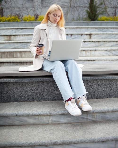 Une jeune étudiante utilise un ordinateur portable et boit du café assise sur un banc