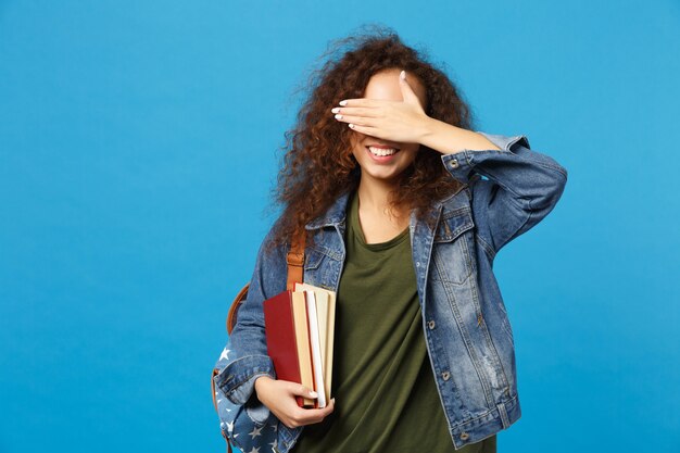 Jeune étudiante en vêtements en jean et sac à dos tient des livres isolés sur un mur bleu