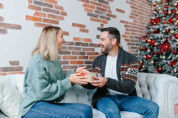 Une jeune famille célèbre ensemble le Nouvel An et Noël, un couple homme et femme se donnent des cadeaux assis sur un canapé à la maison heureux et amoureux