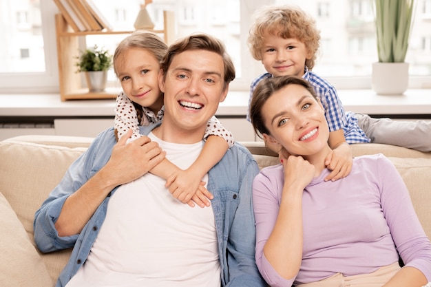 Jeune famille joyeuse de père, mère et deux frères et sœurs mignons embrassant tout en vous relaxant sur un canapé