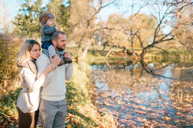 Jeune famille avec leur bébé sur les épaules du père debout près du lac d'automne.