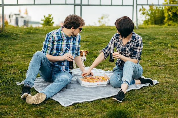 Jeune famille avec leur chien assis sur l'herbe et mangeant une collation de pizza en plein air
