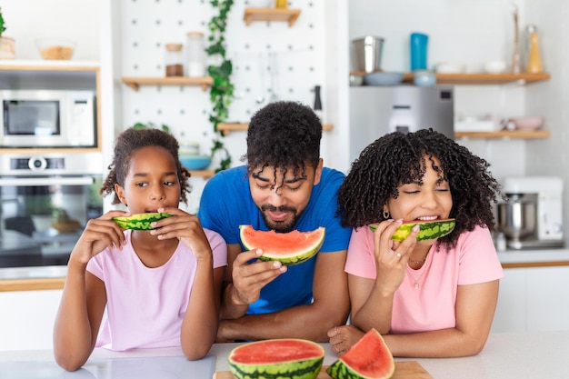 Jeune famille mangeant de la pastèque et s'amusant Famille métisse dans la cuisine mangeant ensemble une tranche de pastèque