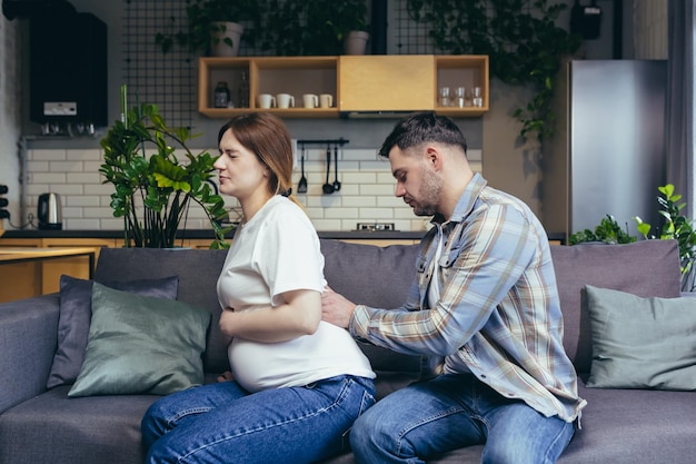 Jeune famille Le mari masse le dos de sa femme enceinte Assise sur le canapé La femme souffre En prévision de l'accouchement